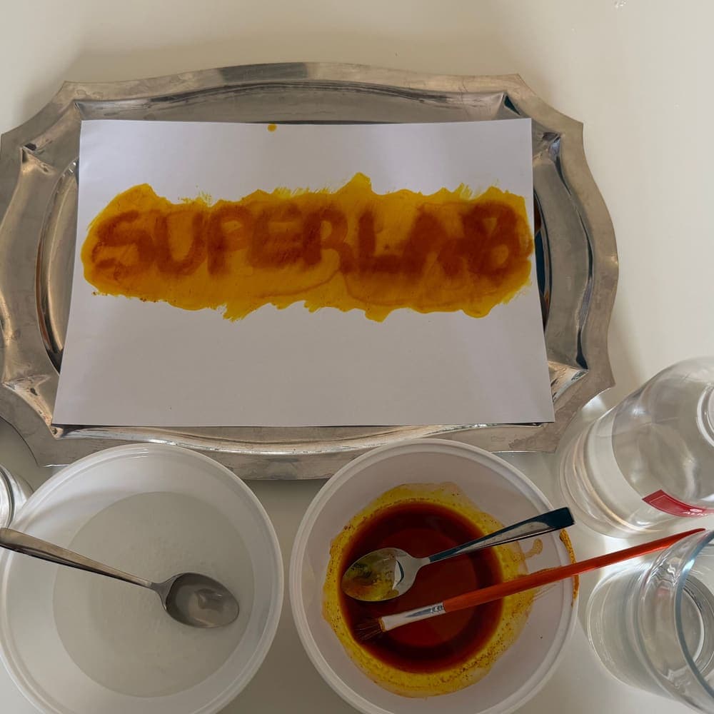 Riječ SUPERLAB napisana kurkumom na papiru, okružena posudama s bojama
